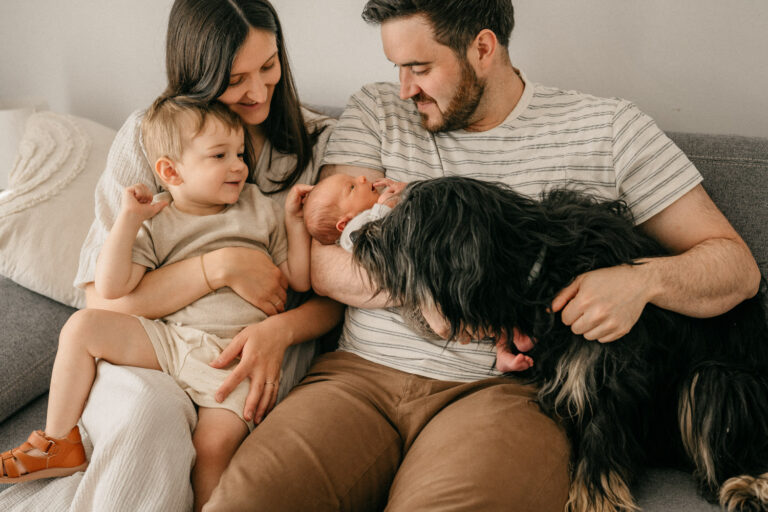 Familie während ihres Newbornshootings. Sie sitzen auf dem Sofa und lachen das Neugeborene an. Der kleine Bruder fasst dem Baby am Köpfchen.