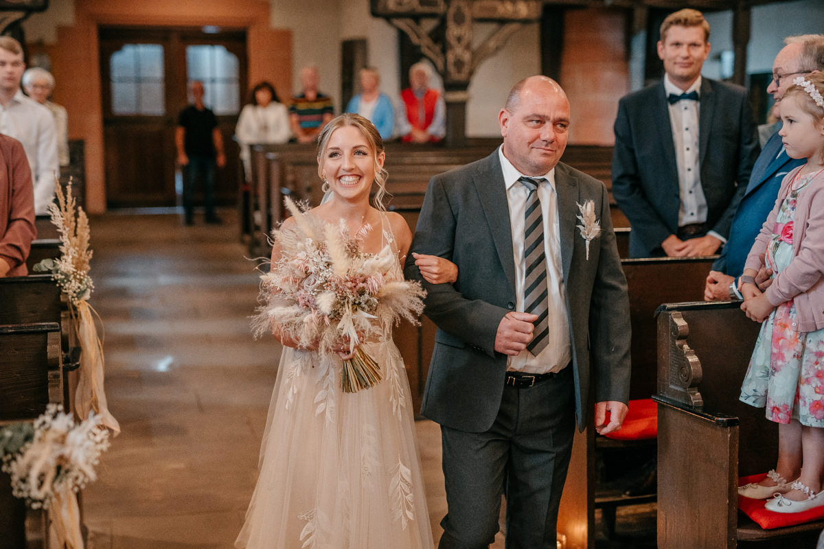 Braut und der Brautvater während des Einzugs in die Kirche. In der Hand hält sie einen Brautstrauß.