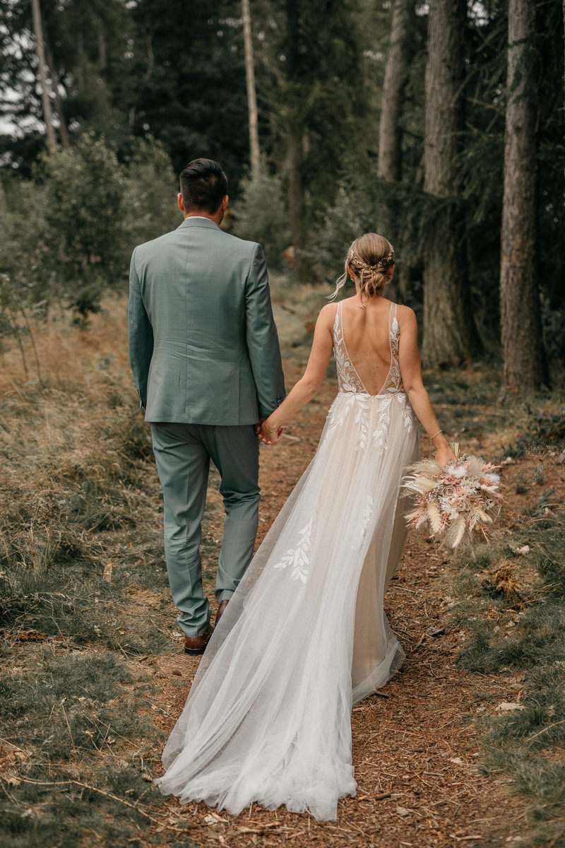 Brautpaar laüft während des Shootings von der Kamera weg. Er trägt einen blau-grünen Anzug mit braunen Schuhen. Sie ein weißes Hochzeitskleid mit floralen Applikationen und einem tiefen Rückenausschnitt. In der Hand hält sie einen Brautstrauß aus Trockenblumen.